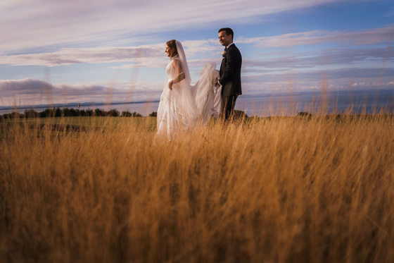Groom holds brides train as they walk through grass together with sea in background